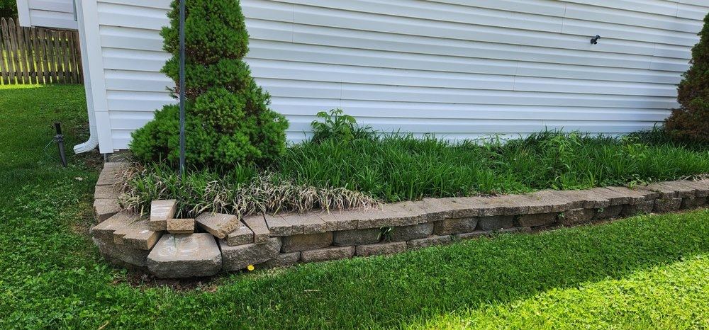 A narrow border along a house wall held by broken, sunken stone edging, with grass growing over it.