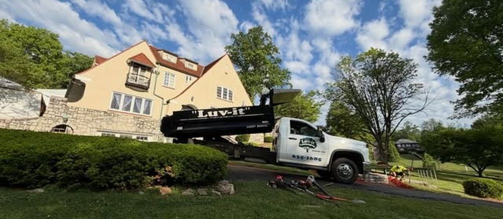 A white pickup and a black dump trailer lettered Luv-It, parked on a driveway in front of a stone and stucco house with tools laid out on the drive.
